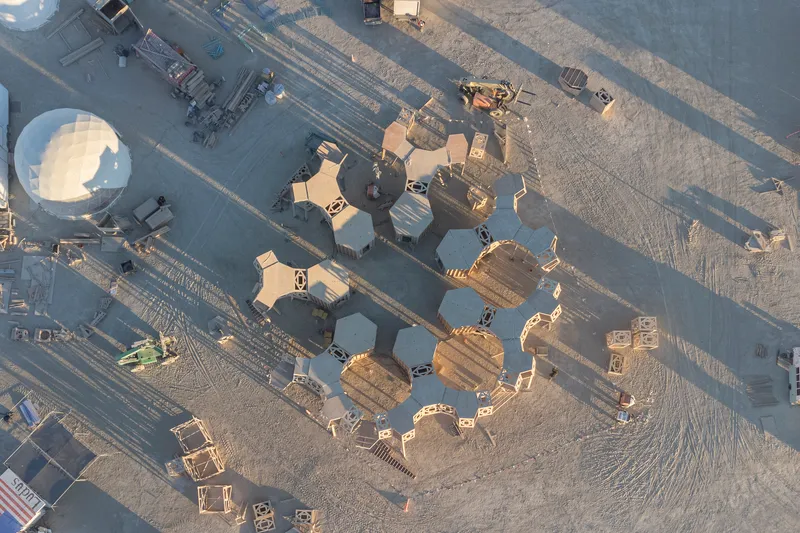 Drone view of hexagonal timber modules clustered on desert playa beside roadway at dawn