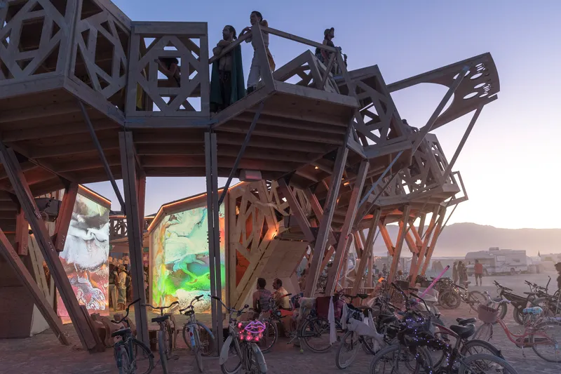 Timber pavilion with open geometric lattice framework and illuminated fabric screens at dusk