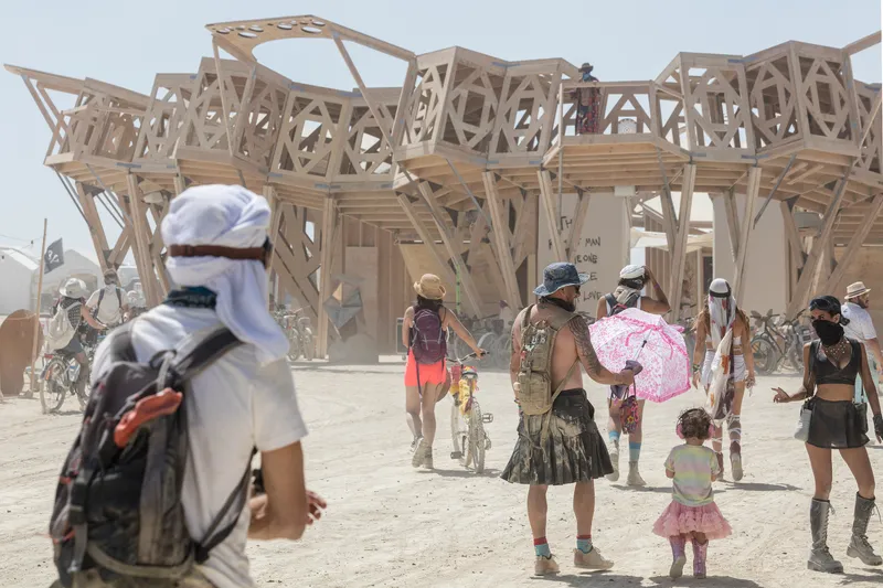 Exterior view of a geometric timber pavilion with latticed walls as visitors walk through dusty conditions
