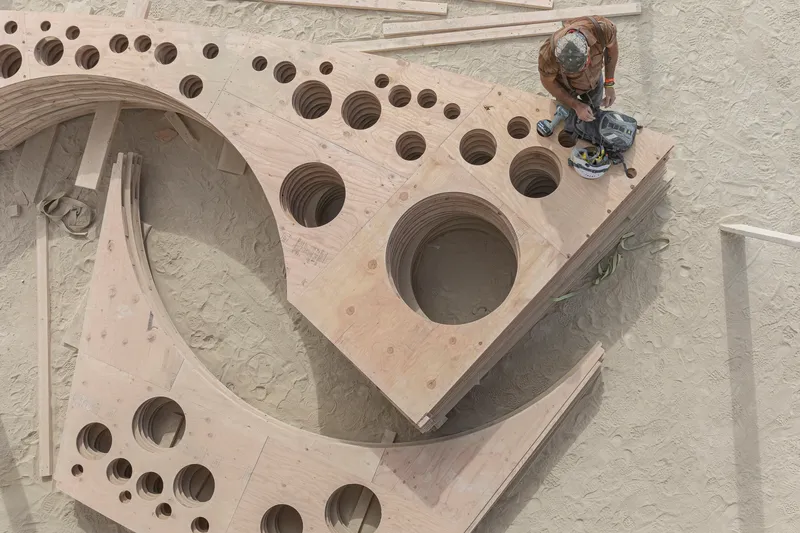 Overhead view of curved plywood panels with circular perforations as a worker applies finish
