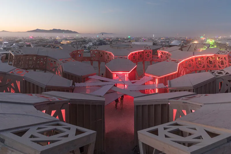 Three-lobed timber structure with red interior lighting at dusk in the desert