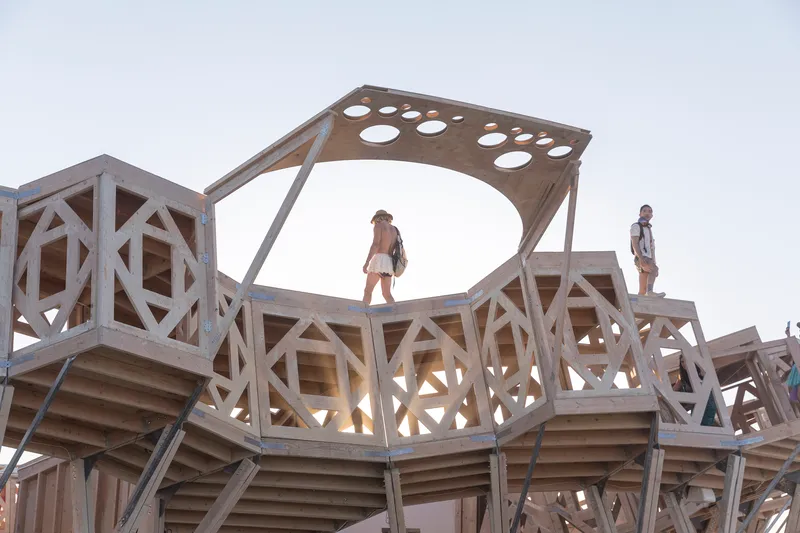 Perforated timber canopy atop geometric wooden framework with visitors silhouetted against the sky