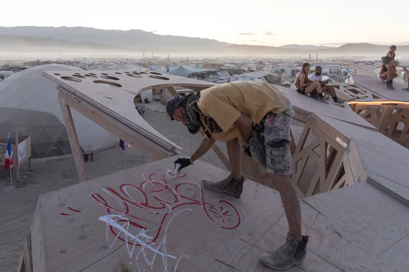 Worker spray-painting graffiti on a plywood panel above the geometric structure at dawn