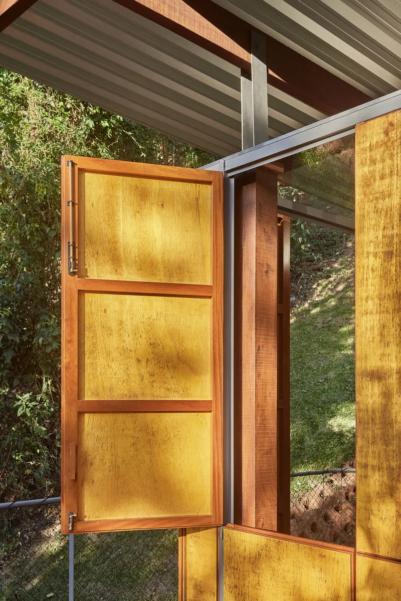 Open plywood shutter revealing the interior and surrounding trees framed by corrugated ceiling and glazing