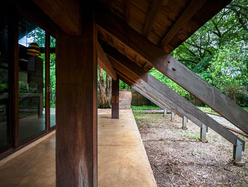 Covered terrace with timber columns and exposed beams extending toward diagonal concrete ramp and trees