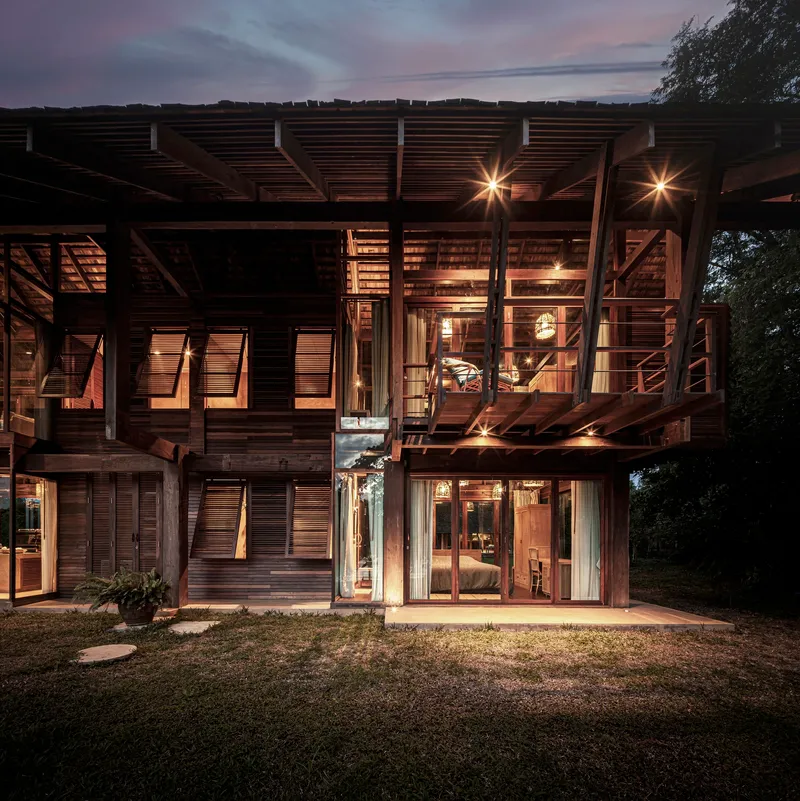 Garden facade illuminated at dusk with layered timber screens and glazed openings beneath exposed structural bracing