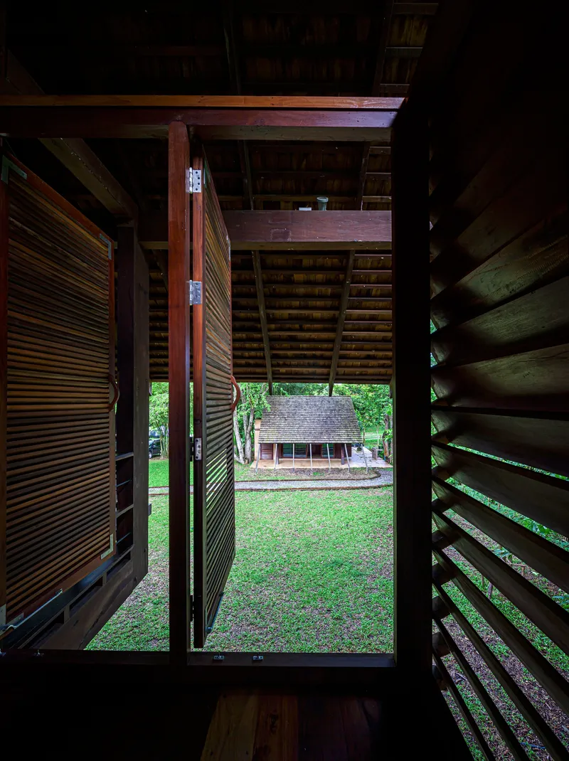 View through open timber louvered doors framing a lawn and pavilion beyond under exposed beam ceiling