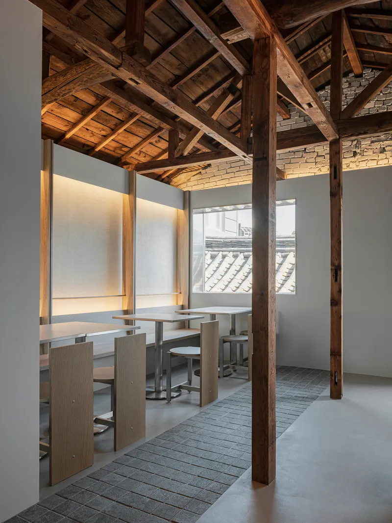 Interior view showing exposed timber roof trusses above a dining area with backlit wall niches and tiled roofs visible through the window