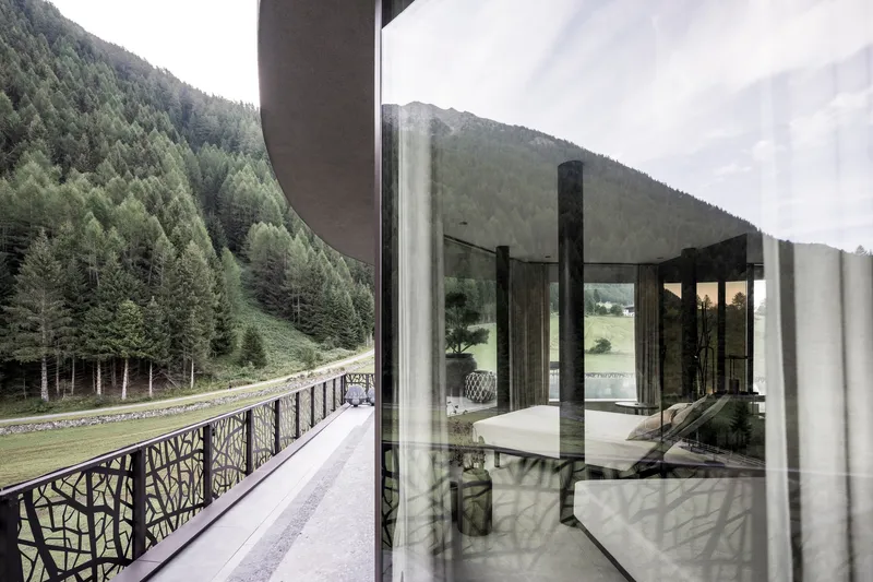 Glass-walled corridor with patterned metal balustrade overlooking a forested mountain slope and meadow