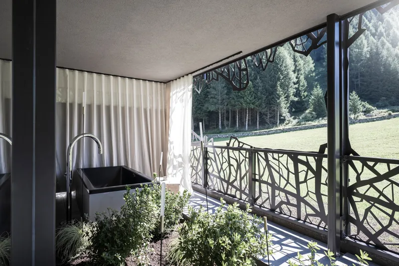 Covered balcony with geometric metal railing, white curtains, and planted greenery facing the forest