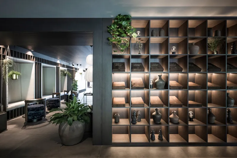 Backlit timber cubby wall displaying ceramic vessels adjacent to an interior courtyard with greenery