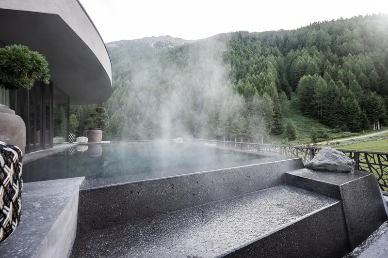Infinity edge pool with dark stone terrazzo finish and rising steam overlooking forested alpine hillside