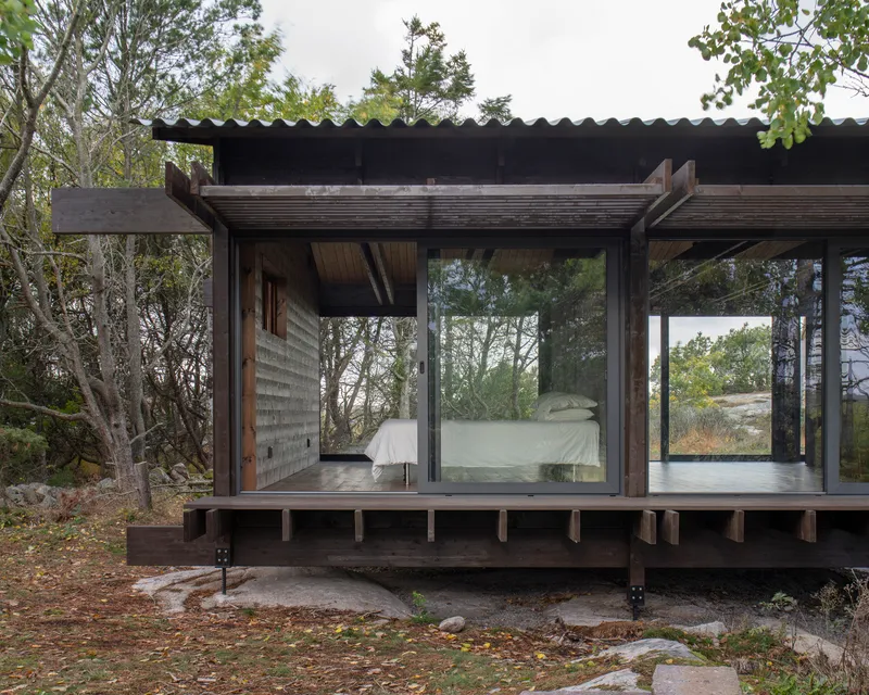 Glass-enclosed bedroom module raised on timber beams with view into surrounding forest on overcast day