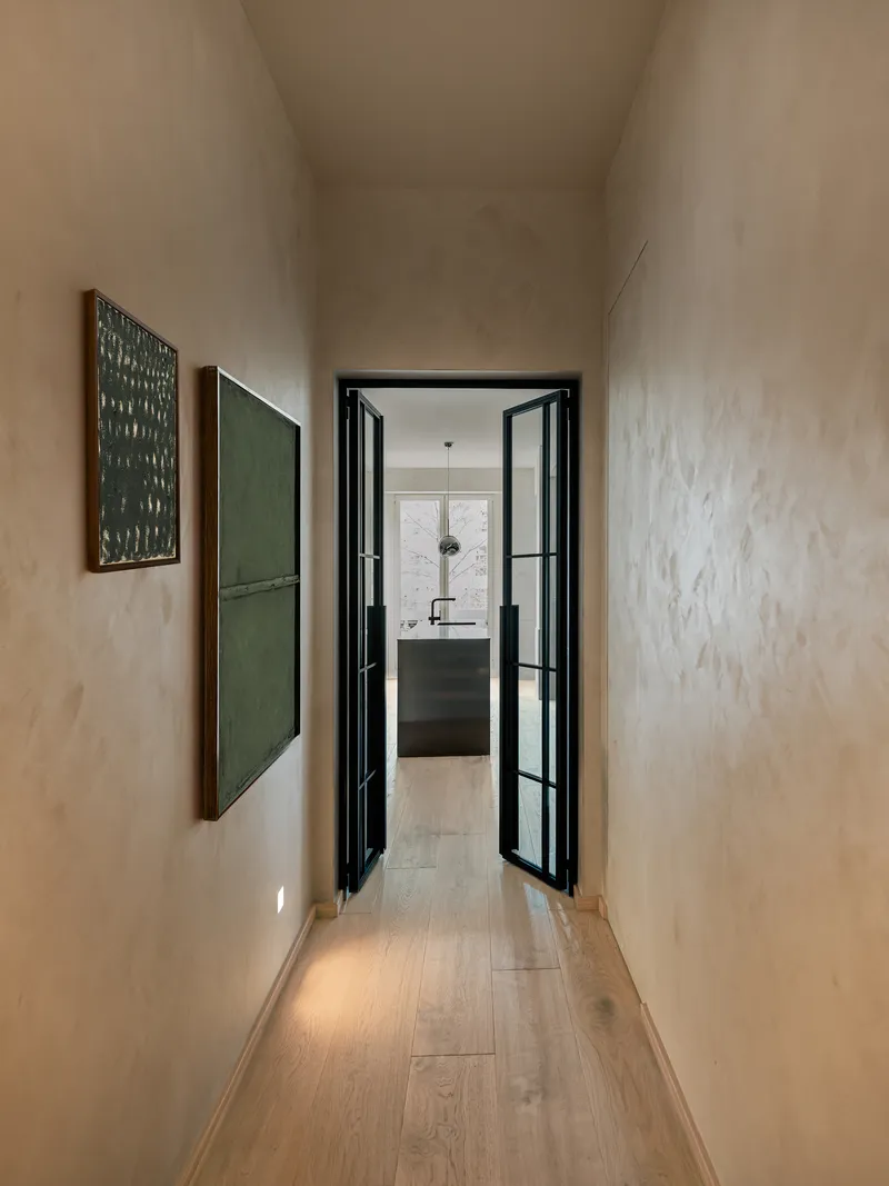 Narrow corridor with limewashed plaster walls leading to the kitchen through black-framed glass doors