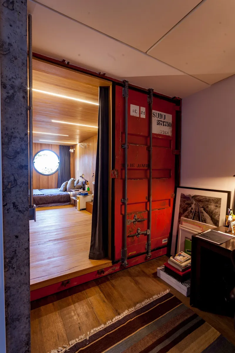 Repurposed red shipping container door on rails opening to a timber-clad bedroom