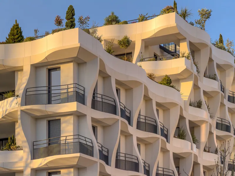 Undulating white facade with recessed balconies and rooftop plantings in afternoon sunlight