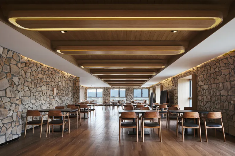 Long dining hall with illuminated stone perimeter walls, stepped timber ceiling, and walnut flooring under daylight