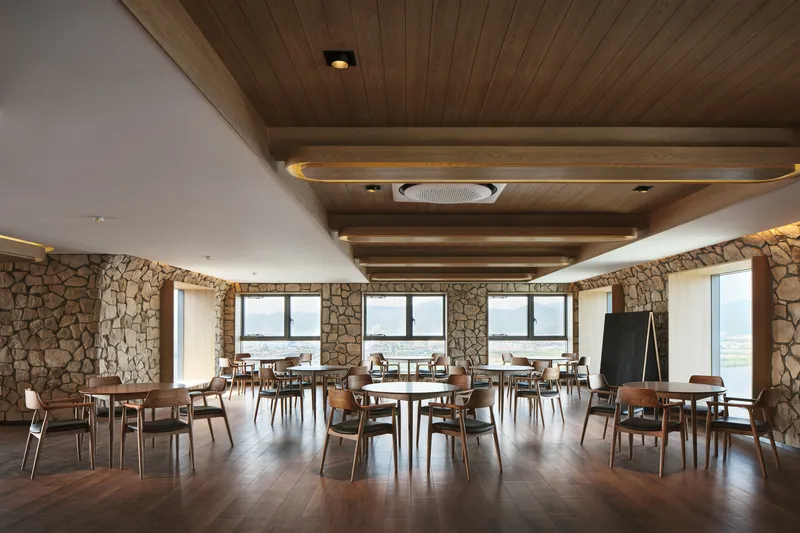 Dining hall with timber-slatted ceiling above white plaster volumes and fieldstone walls with river views