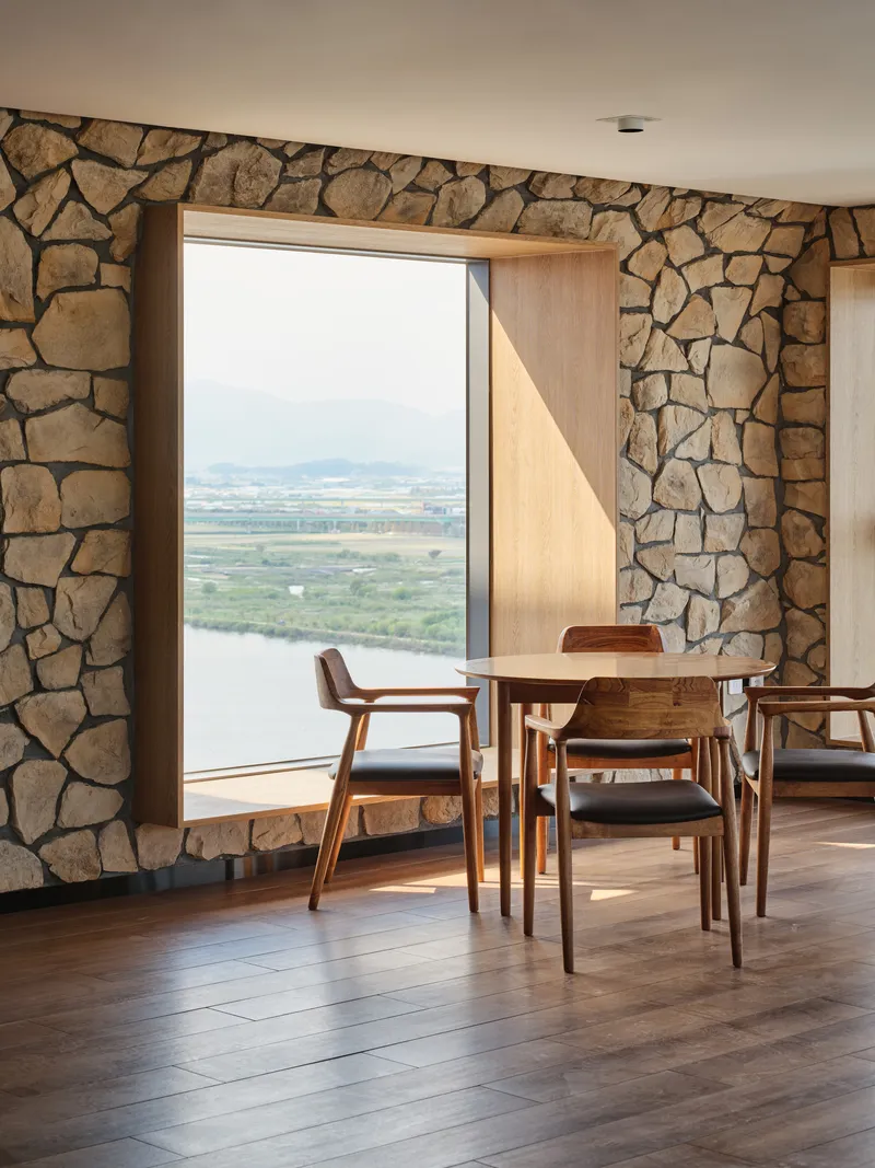 Dining area with timber-framed window opening onto valley and lake view through rough stone wall