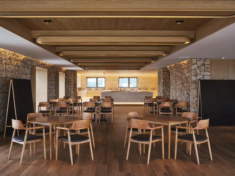 Dining area with recessed timber ceiling panels, backlit stone walls, and rows of oak tables and chairs