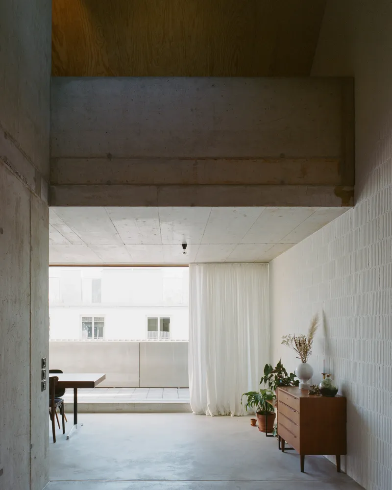 Open-plan interior with exposed concrete beam, polished floor and a wooden sideboard with potted plants near sheer curtains