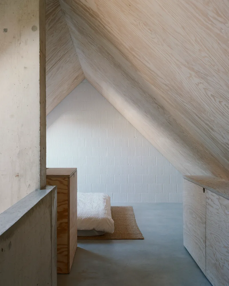 Attic space with diagonal wood-clad ceiling, concrete walls and a cushion on woven mat under soft daylight