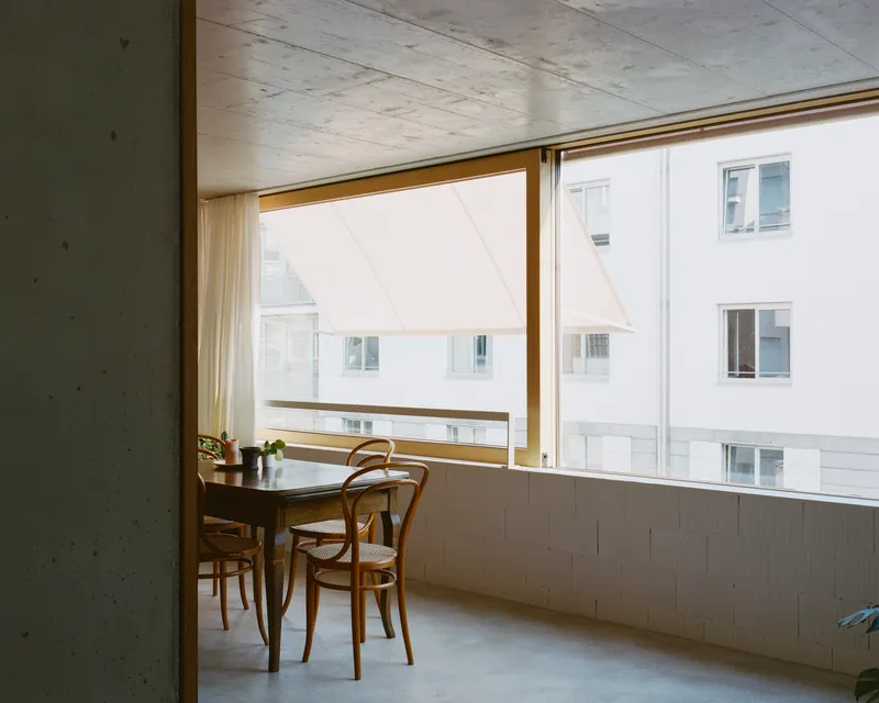 Interior corner with exposed concrete ceiling, wood-framed window and bentwood chairs at a table overlooking white facades