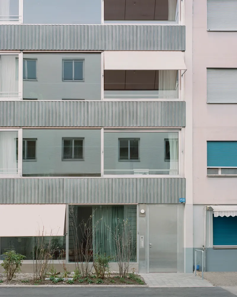 Street facade with corrugated metal balconies, tiled panels and planted shrubs at ground level in overcast light