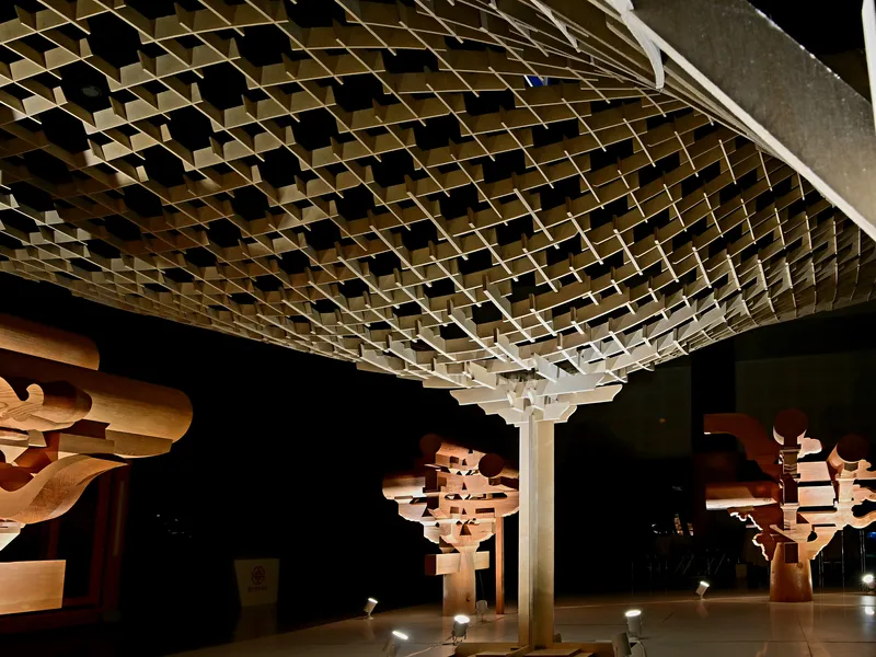 Undulating timber canopy suspended from a central column in an exhibition space at night