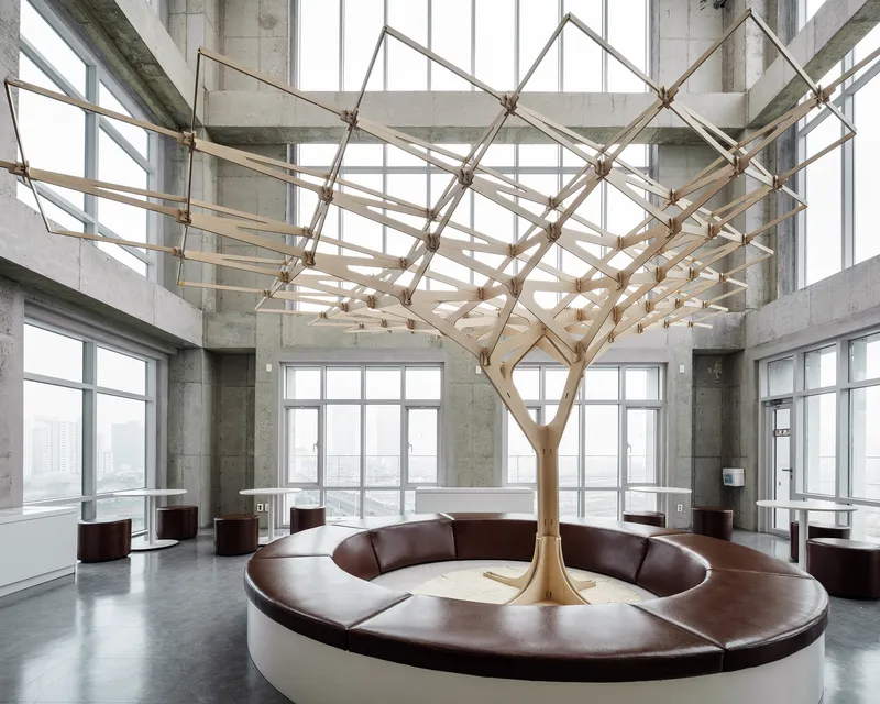 Tree-like timber canopy with branching lattice structure above a circular bench in a concrete-framed room with floor-to-ceiling windows