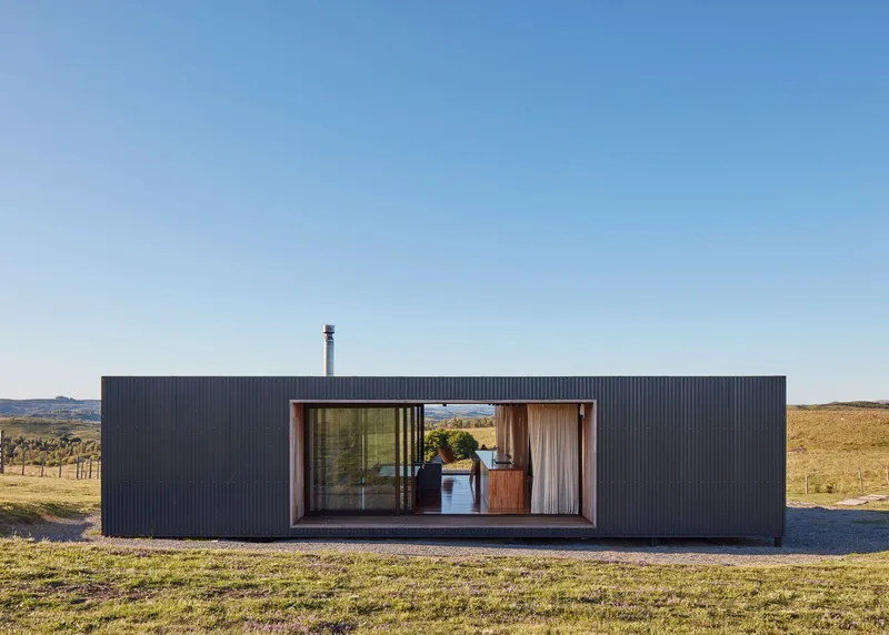 Front elevation showing corrugated metal cladding with central timber-framed glazed opening onto gravel terrace