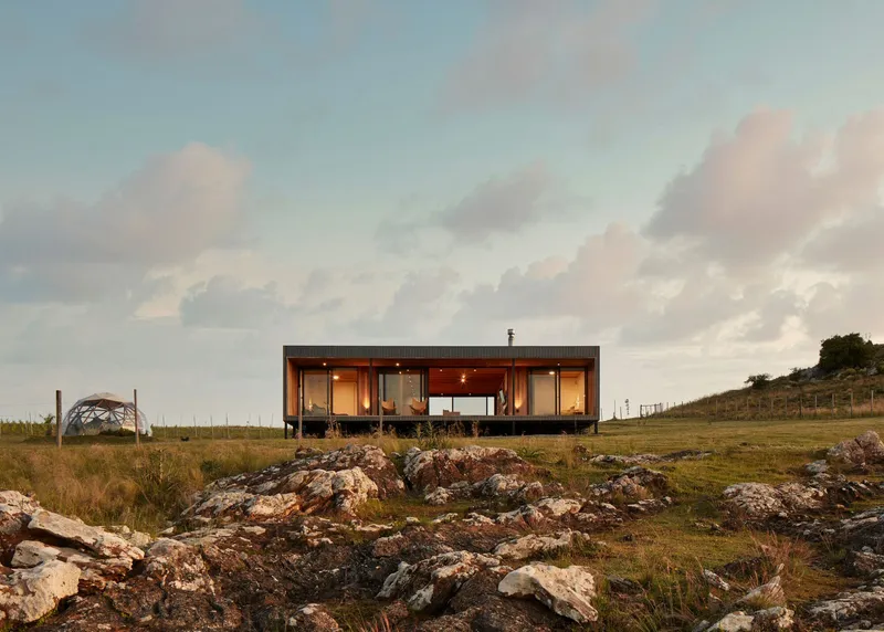Glass-walled volume on rocky terrain illuminated at twilight beneath a cloudy sky