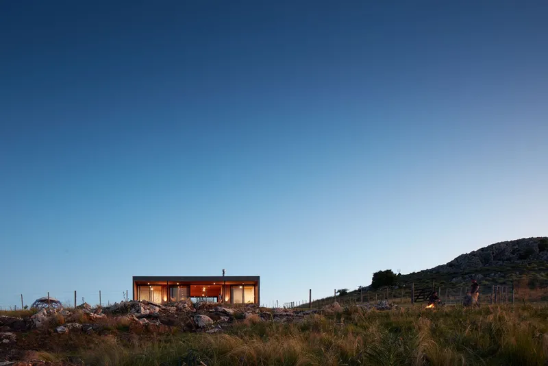 Oblique view across native grasses and limestone boulders toward the glowing structure at dusk