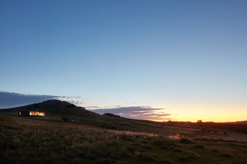 Wide landscape view of the illuminated residence against a hillside and colorful sunset horizon