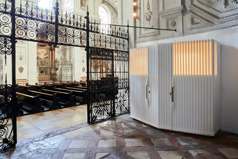 White fluted timber confessional booths positioned beside ornate black wrought iron gates and historic patterned stone flooring