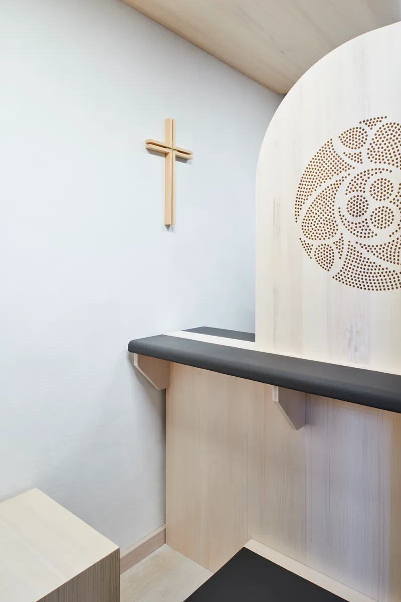 Interior confessional space with pale timber bench, simple timber cross on white wall, and perforated pattern detail