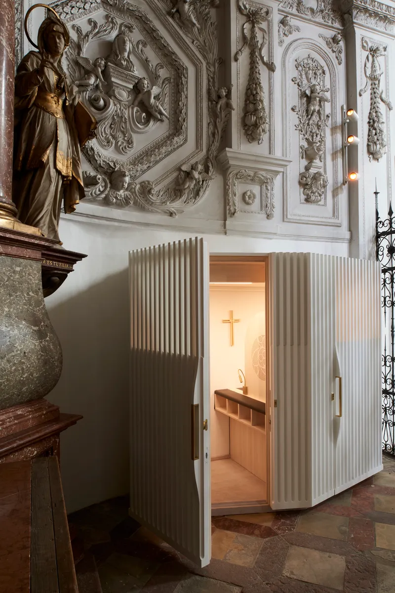 Open doorway in the ribbed timber enclosure revealing the lit confessional interior with cross and seating