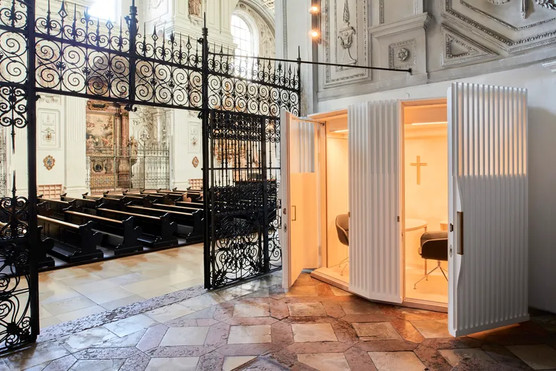 White fluted confessional booth inserted within a historic church interior behind ornamental wrought iron gates