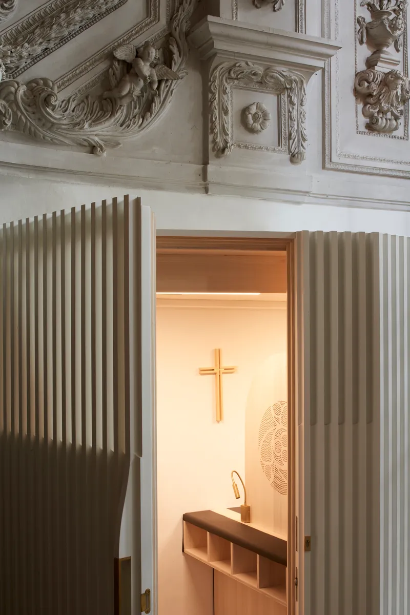 Open doorway of the white fluted confessional revealing warm interior light beneath ornate ceiling plasterwork
