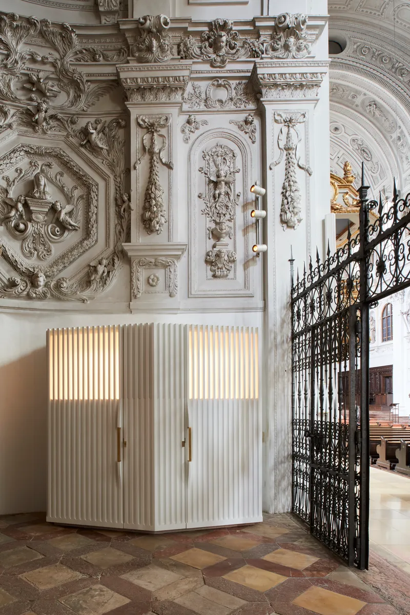 Corner view of the white fluted confessional with backlit panels against the ornately carved church wall