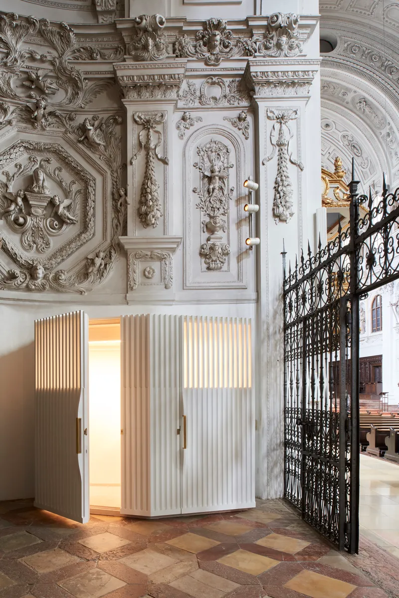 White fluted confessional booth with open door showing illuminated interior and brass handles in the church nave