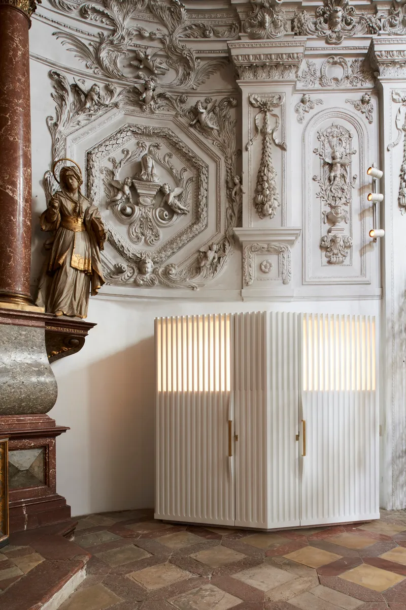 White fluted confessional with backlit panels positioned beneath decorative plasterwork and beside a marble column