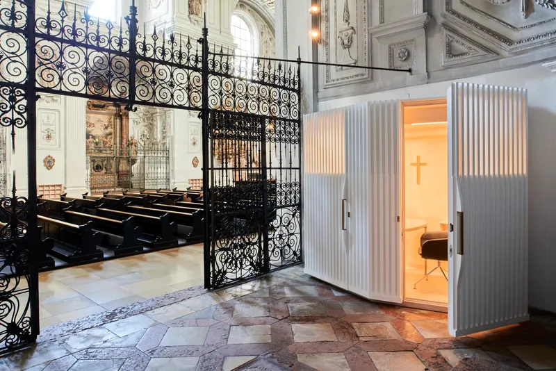White confessional booth with illuminated cross and vertical fins beside ornate wrought iron gates and pews