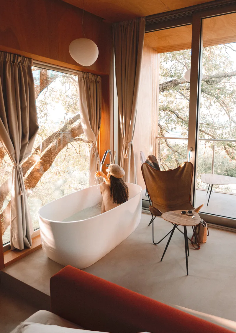 Freestanding bathtub beside floor-to-ceiling windows with a person bathing in morning light