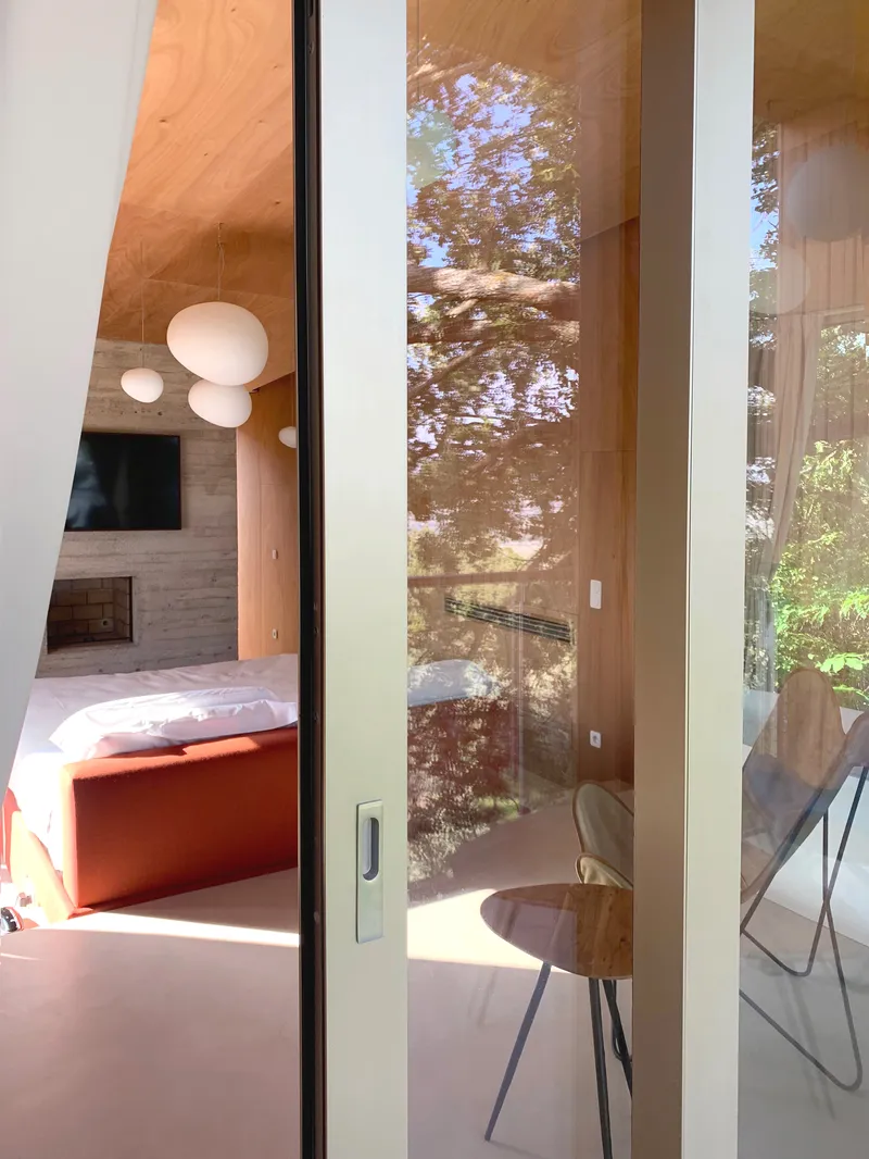 Glass door reflecting trees and revealing bedroom with plywood ceiling beyond