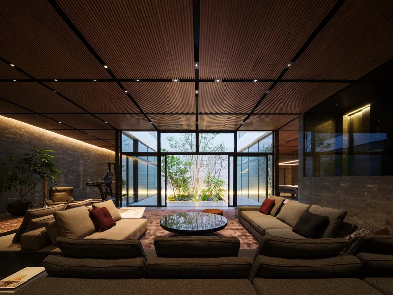 Seating area beneath gridded timber ceiling with full-height glazed walls framing courtyard garden at dusk