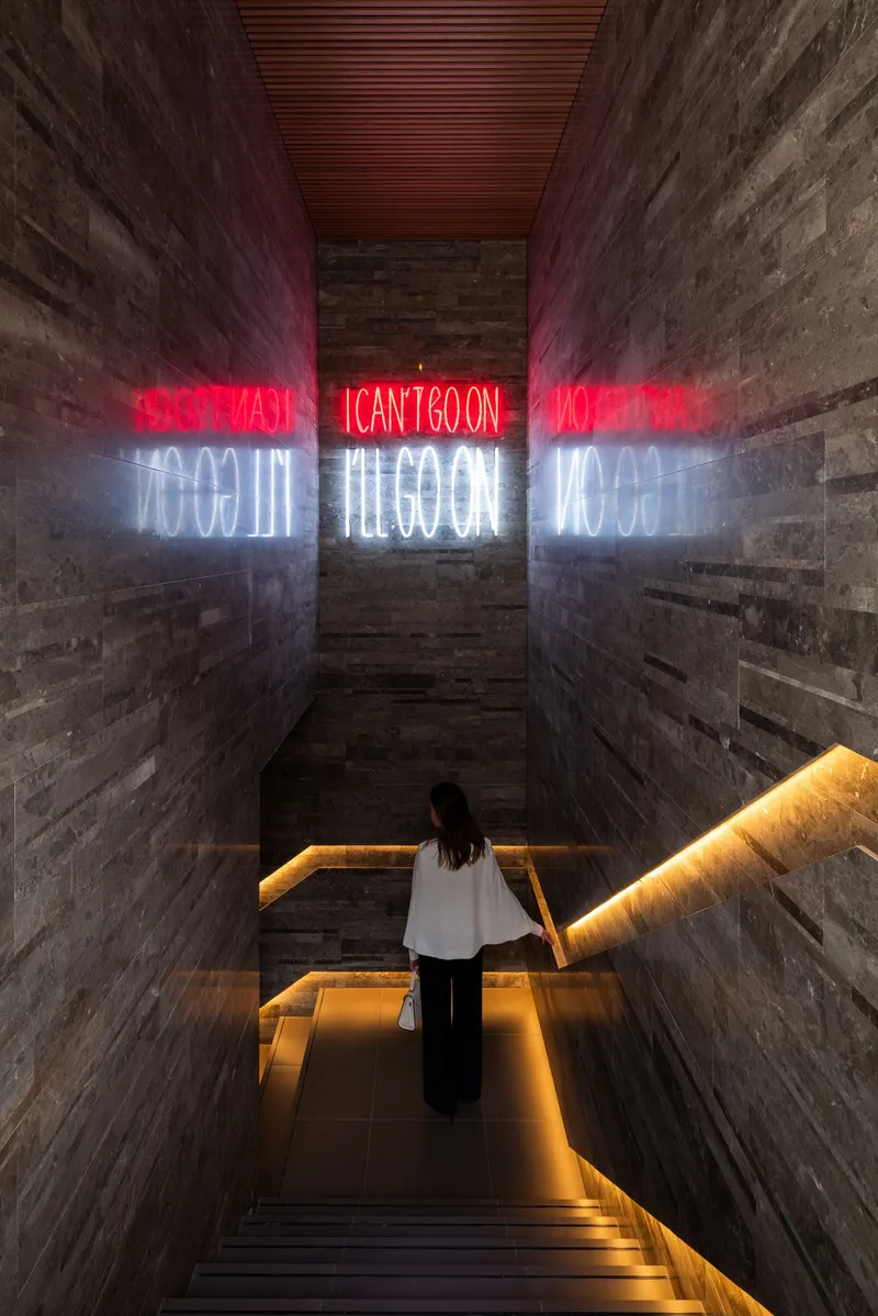 View down a concrete stairwell with red and white neon text reflected on board-formed walls