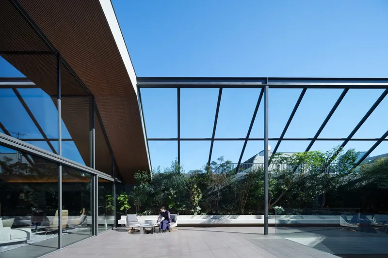 Covered terrace with steel frame and glass roof looking toward planted screening and loungers