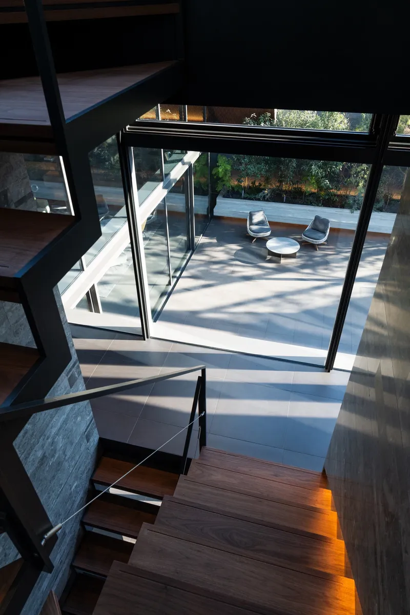 View down timber staircase through slanted glazing to paved terrace with seating below