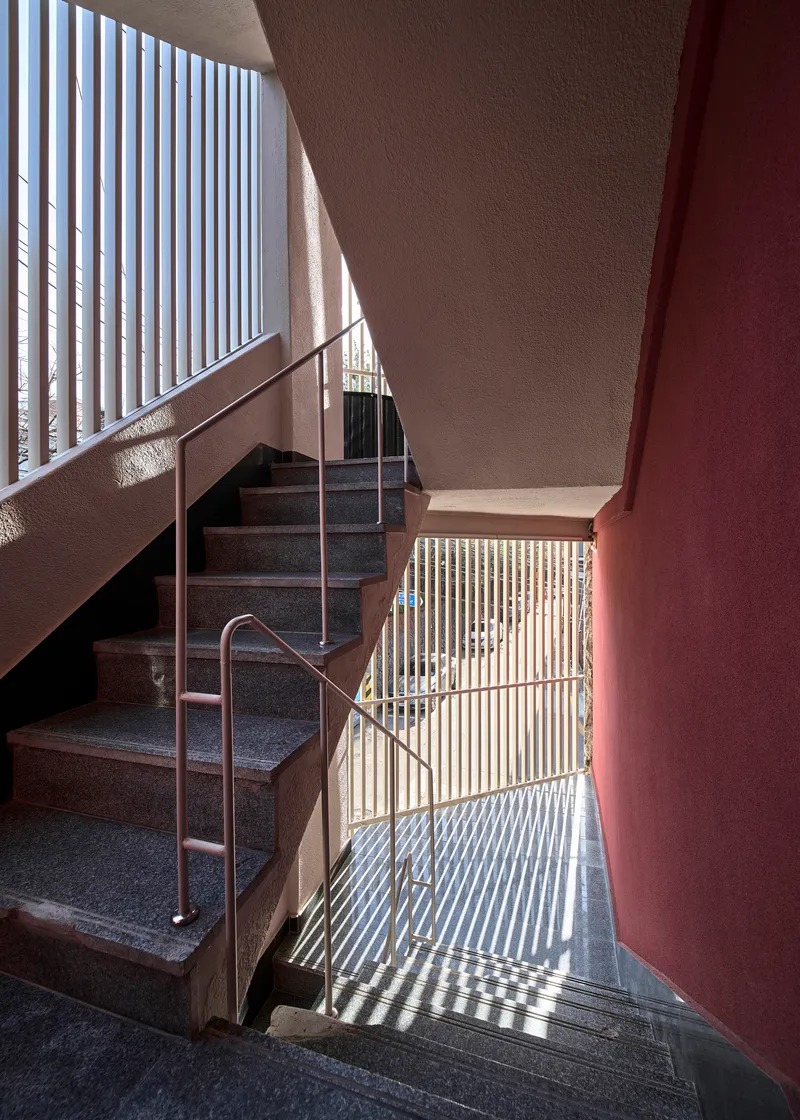Stairwell with vertical slat screens casting striped shadows across terrazzo steps and maroon walls